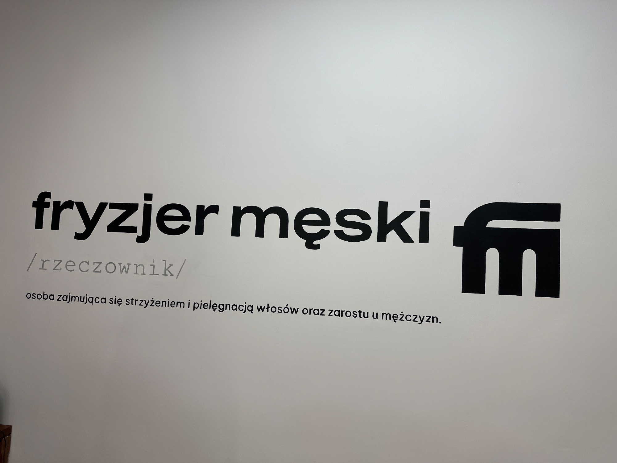 Napis „fryzjer męski /rzeczownik/” z definicją i znakiem fm na białej ścianie salonu